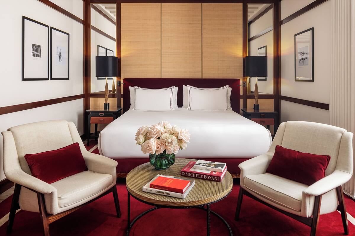 A hotel room with a large bed, two armchairs, a small round table with a vase of flowers and books, and framed pictures on the walls.