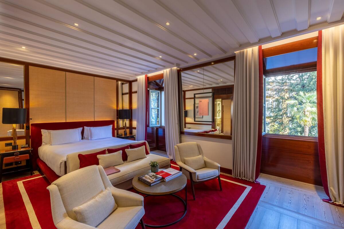 A hotel room with a large bed, a sofa, a chair, a small table, and a window with curtains.