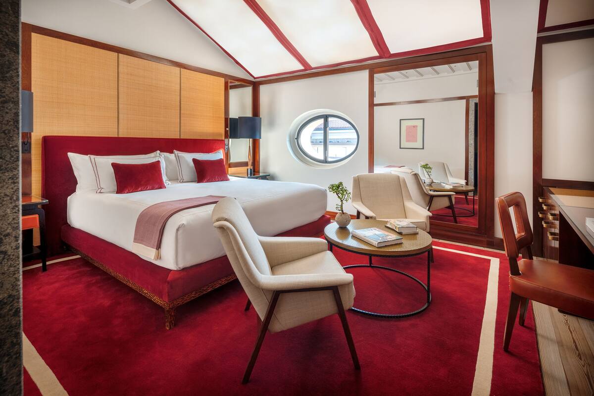 A hotel room with a large bed, a sitting area with a table and chairs, a round window, and a view of the outside.