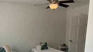 Habitación con dos camas individuales, cada una con cubrecolchón verde y ropa de cama blanca. Hay un ventilador de techo con una lámpara sobre las camas, y una puerta blanca a la derecha.