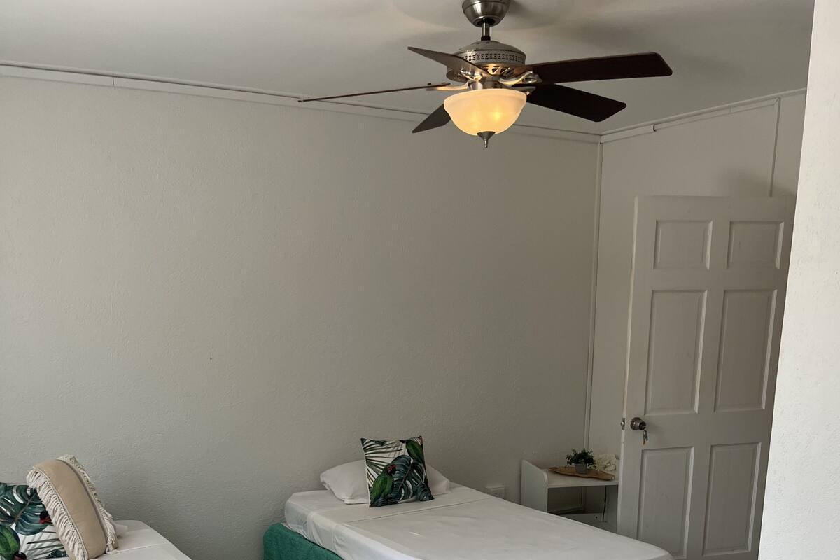 Habitación con dos camas individuales, cada una con cubrecolchón verde y ropa de cama blanca. Hay un ventilador de techo con una lámpara sobre las camas, y una puerta blanca a la derecha.