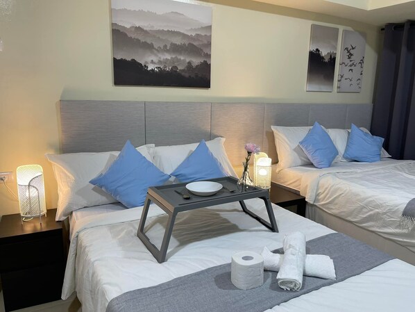 A hotel room with two beds, each with white bedding and blue pillows. There is a tray table with a bowl, a vase with a flower, and a roll of toilet paper on the bed.