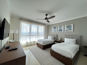Habitación de hotel con dos camas, un escritorio, televisión, ventilador de techo y obras de arte en la pared.