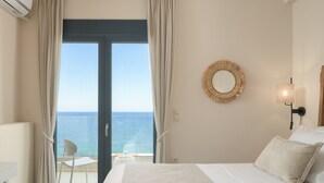 Hotelkamer met een bed, nachtkastje, stoel, uitzicht op zee door een glazen deur en een ronde spiegel aan de muur.