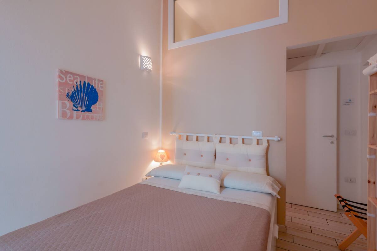 Ein Schlafzimmer mit Bett, Nachttisch, Wandlampe, einer Tür und einer Bank.