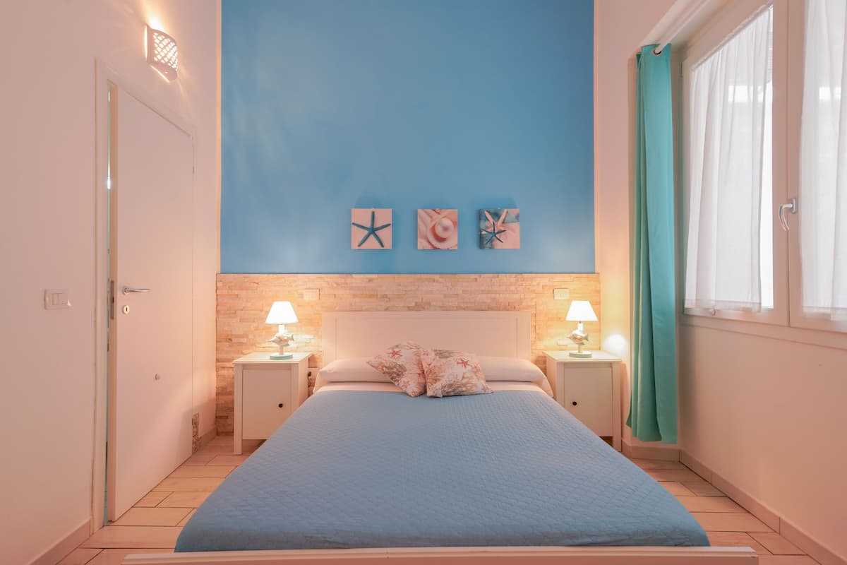 Ein Schlafzimmer mit einem Bett, zwei Nachttischen mit Lampen, einem Fenster mit Vorhang und drei Wandbildern.