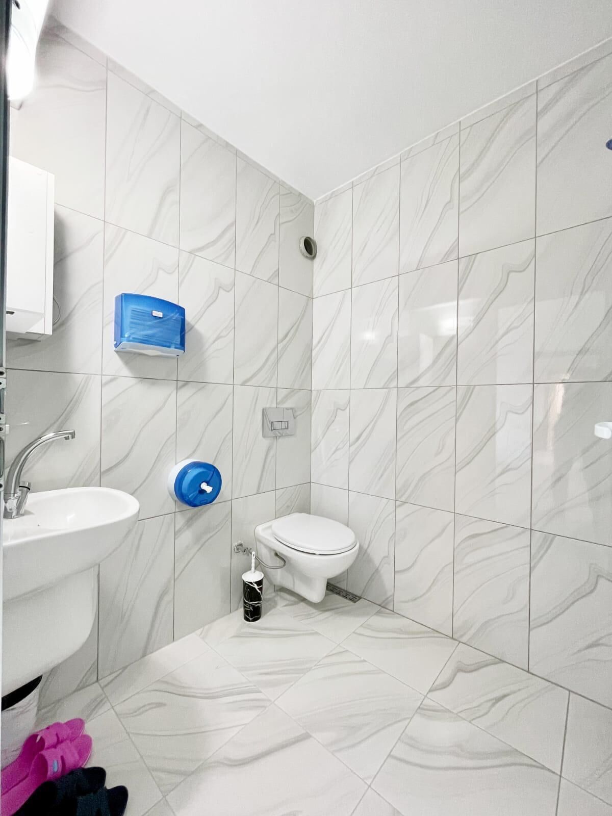 Un baño con baldosas de mármol blanco, un lavabo blanco, un inodoro y un dispensador de jabón azul.