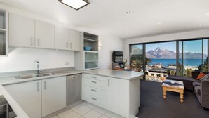 A modern kitchen with white cabinets and a built-in dishwasher. There is a dining area with a table and chairs, and a large window offering a view of a mountainous landscape and a body of water. - Welcome to Ballarat Bungalow. A Chic Central Stay with Easy Access to Queenstown’s Best! (Queenstown)