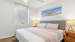 A bedroom with a bed, a window with blinds, a painting of a beach, and bedside lamps. - Aquaview Unit 304, 29 Canberra Tce, Kings Beach (Kings Beach)