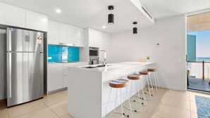 A modern kitchen with a stainless steel refrigerator, a blue backsplash, white cabinetry, and a bar area with stools. - Aquaview Unit 304, 29 Canberra Tce, Kings Beach (Kings Beach)
