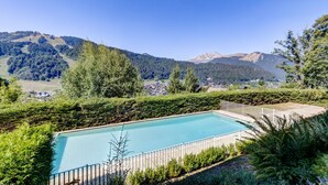 A rectangular swimming pool with a white fence, surrounded by lush greenery and a mountainous backdrop. - Les Marmottes du Morzena (Morzine)