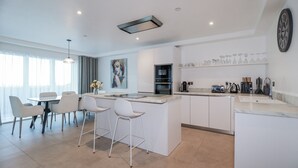 A modern kitchen with a central island, white cabinetry, and a dining area with chairs. - 13 Cliff Edge (Newquay)