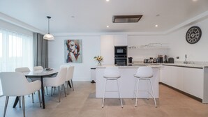 A modern kitchen with white cabinetry, a central island with bar stools, and a dining area with white chairs. - 13 Cliff Edge (Newquay)