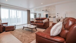 A spacious living room with leather sofas, a coffee table, and a large window with curtains. - 13 Cliff Edge (Newquay)