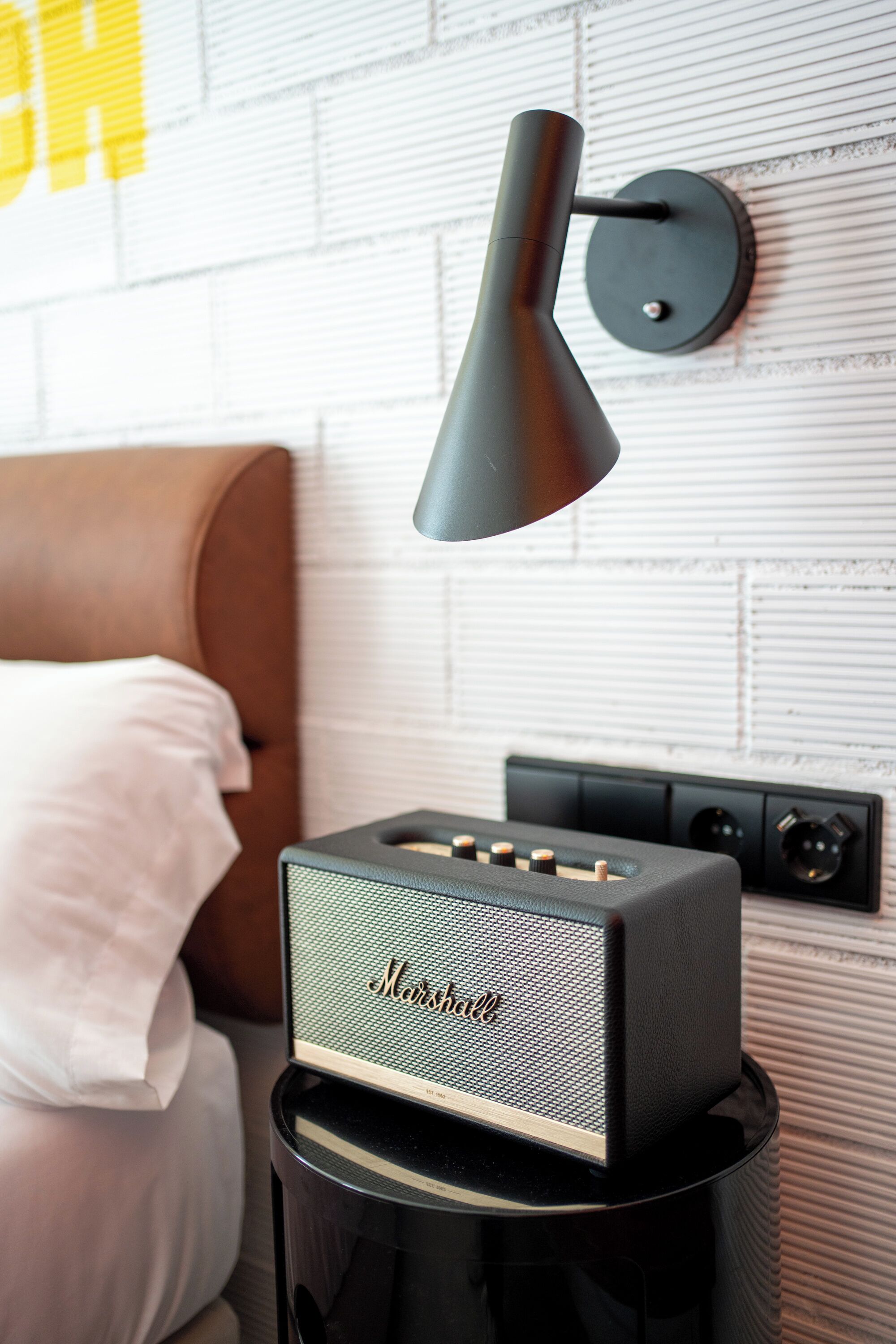 A bed with a pillow, a wall-mounted lamp, and a speaker on a stand.
