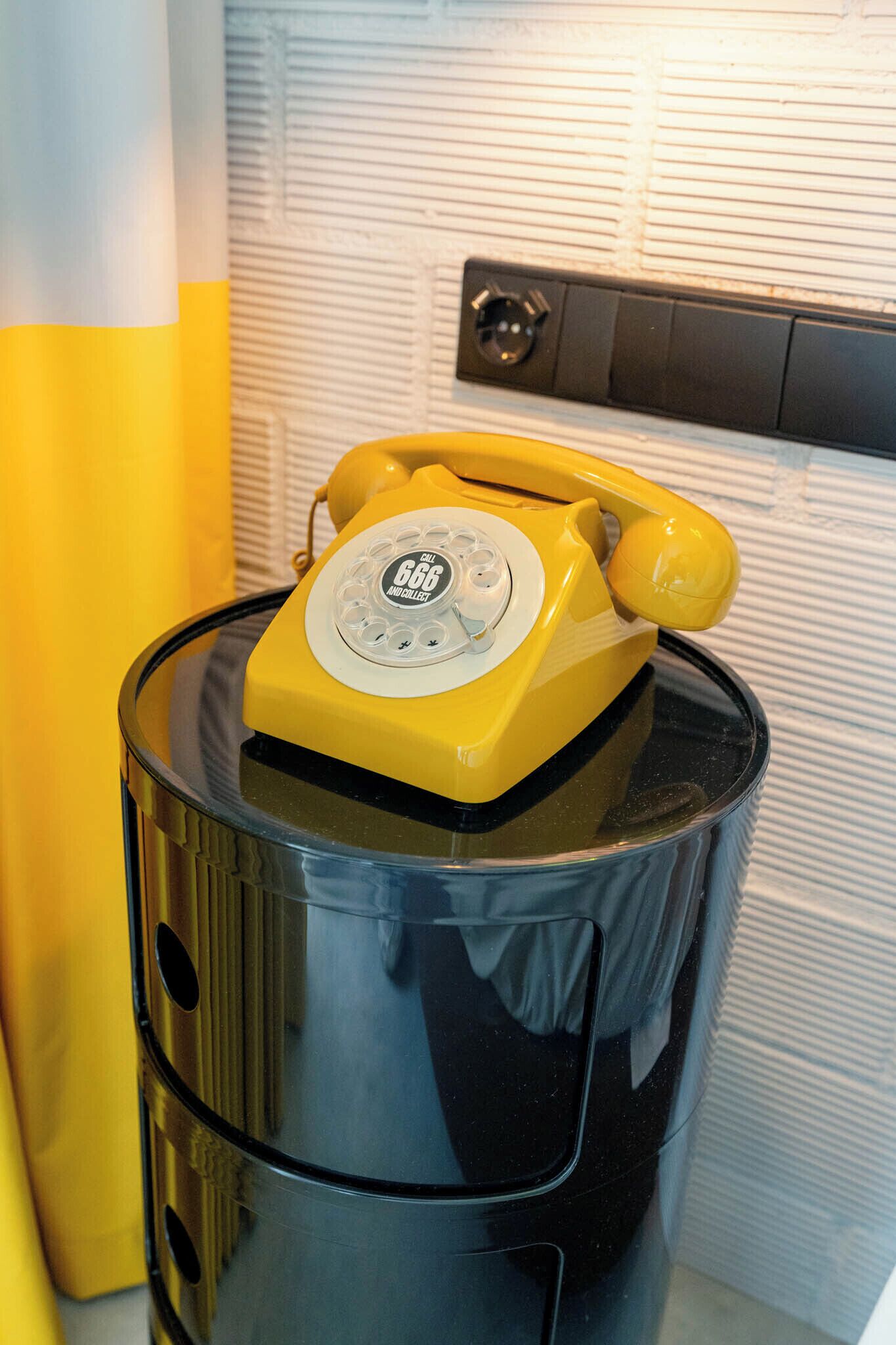 A yellow rotary phone on a black cylindrical object, possibly a speaker or a container, with a textured wall in the background.