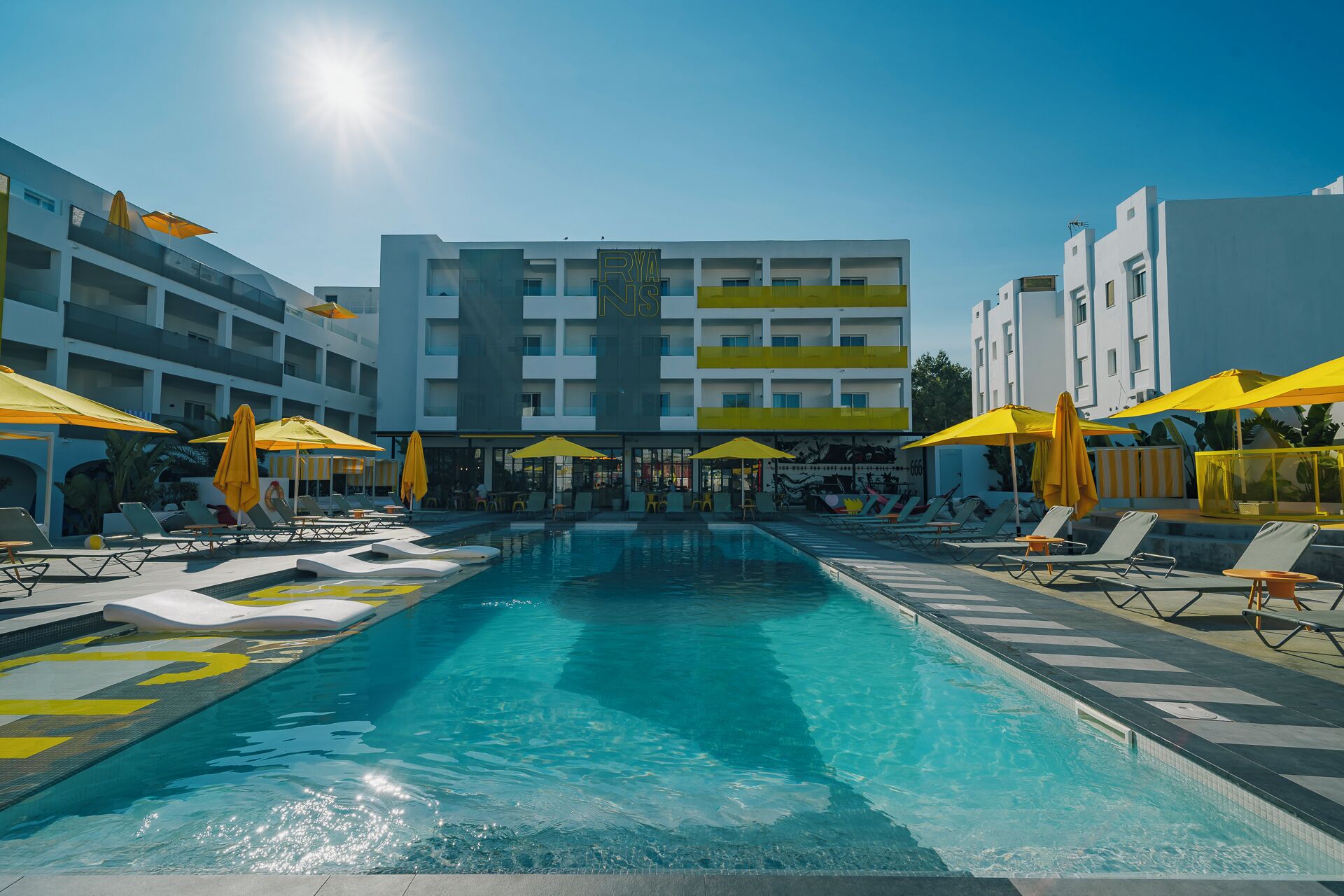 A swimming pool with sun loungers and umbrellas, surrounded by a building with balconies.
