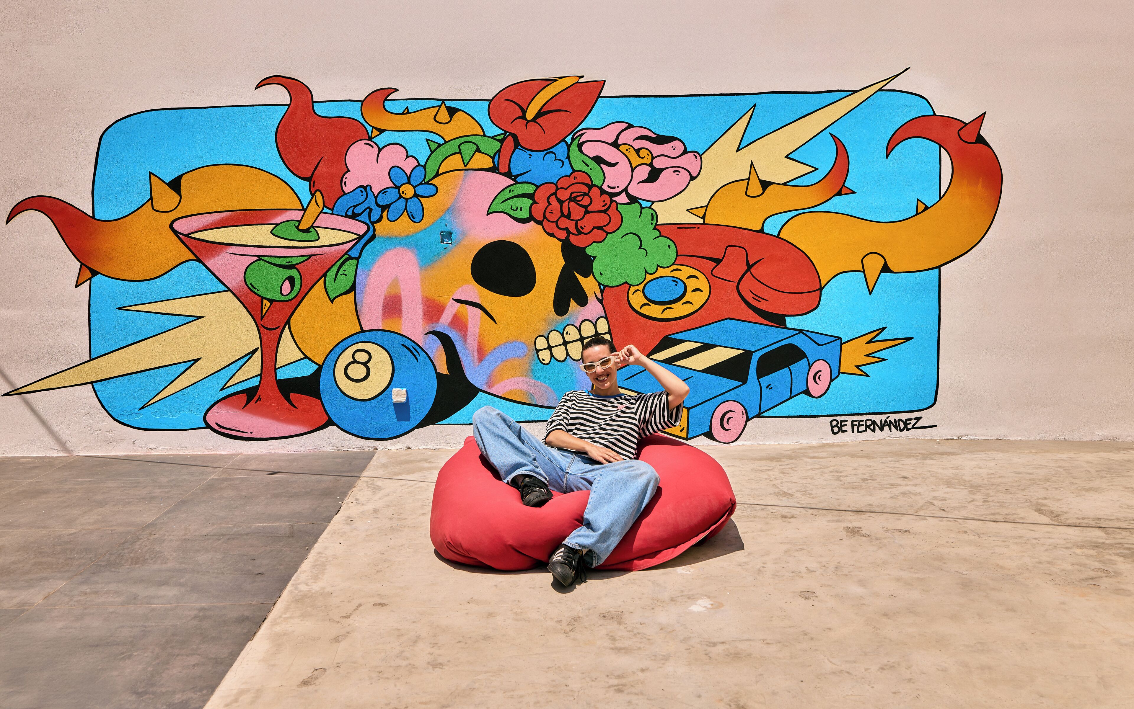 A person sitting on a red bean bag chair in front of a colorful mural with a skull, flowers, and a car.