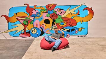A person sitting on a red bean bag chair in front of a colorful mural with a skull, flowers, and a car.