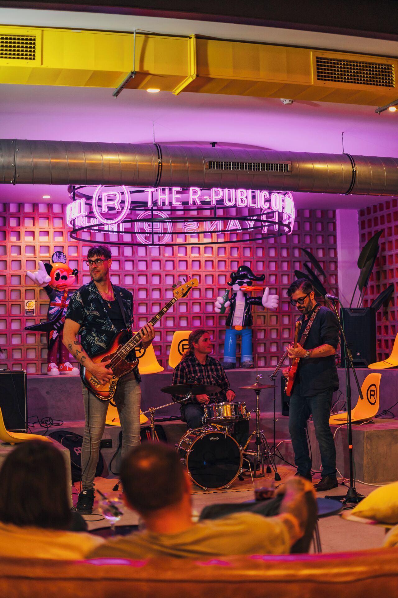 A band performing on a stage with a backdrop featuring a grid pattern and a neon sign.