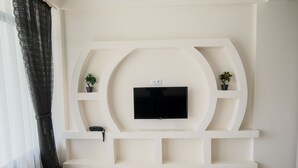 A modern living room with a flat-screen TV mounted on a white wall, flanked by two small shelves with potted plants. - Merkür Hotel (Ihsaniye)
