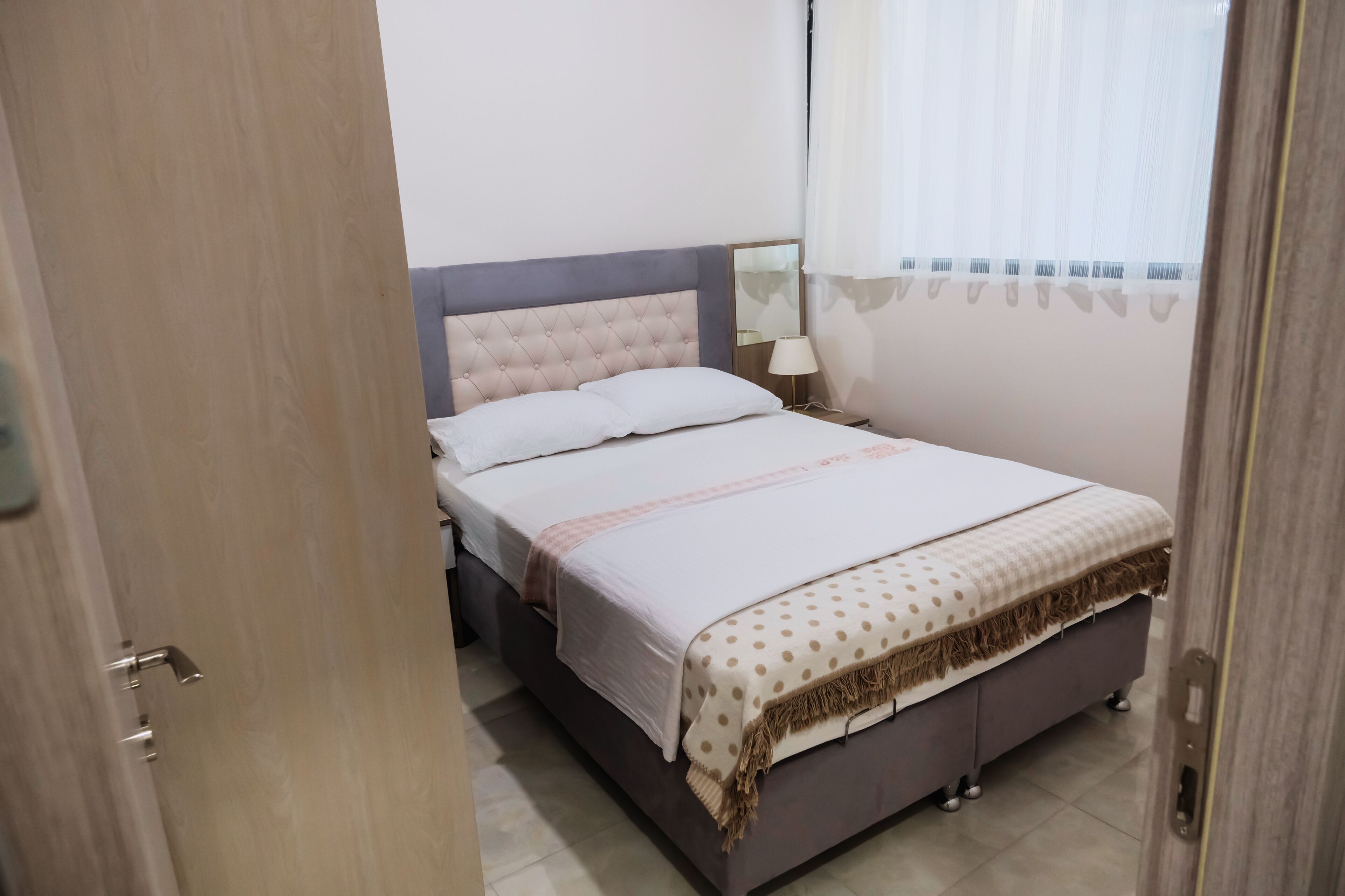 A neatly made bed with a tufted headboard, a bedside lamp, and a window with sheer curtains.
