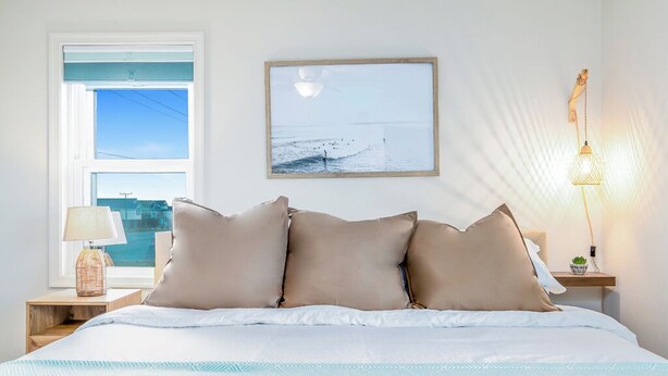 Una cama bien hecha con una colcha estampada, un cuadro con una escena de playa y una lámpara de noche en una mesita.