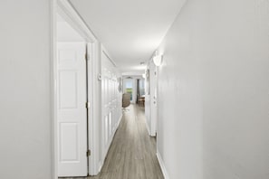 A hallway with white walls and wooden flooring, leading to a dining area with chairs and a table.