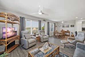 Un salon spacieux avec un téléviseur, deux fauteuils gris, une table à manger avec des chaises et un ventilateur de plafond.