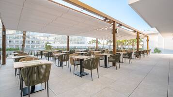 Un área de comedor al aire libre con varias mesas y sillas, piso de baldosas y vista a un edificio al fondo.