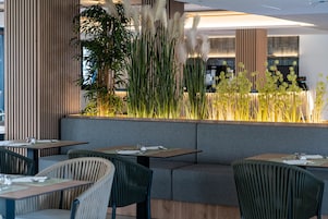 Un restaurante moderno con una zona de estar que cuenta con bancos acolchados y mesas, rodeado de plantas altas y hierbas decorativas.
