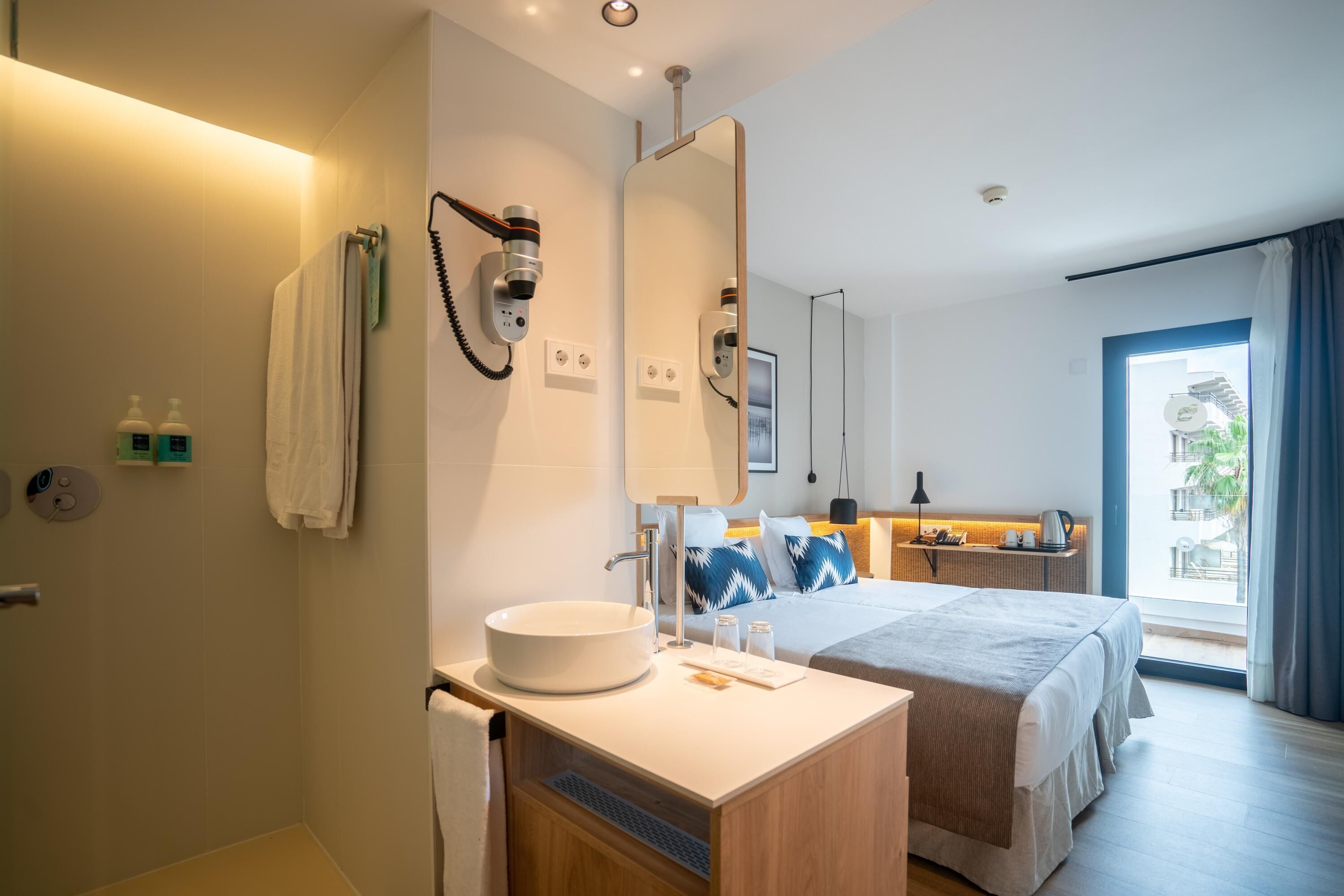 Une chambre d’hôtel moderne avec un grand lit, une table de chevet avec un miroir, un lavabo rond et un espace douche.