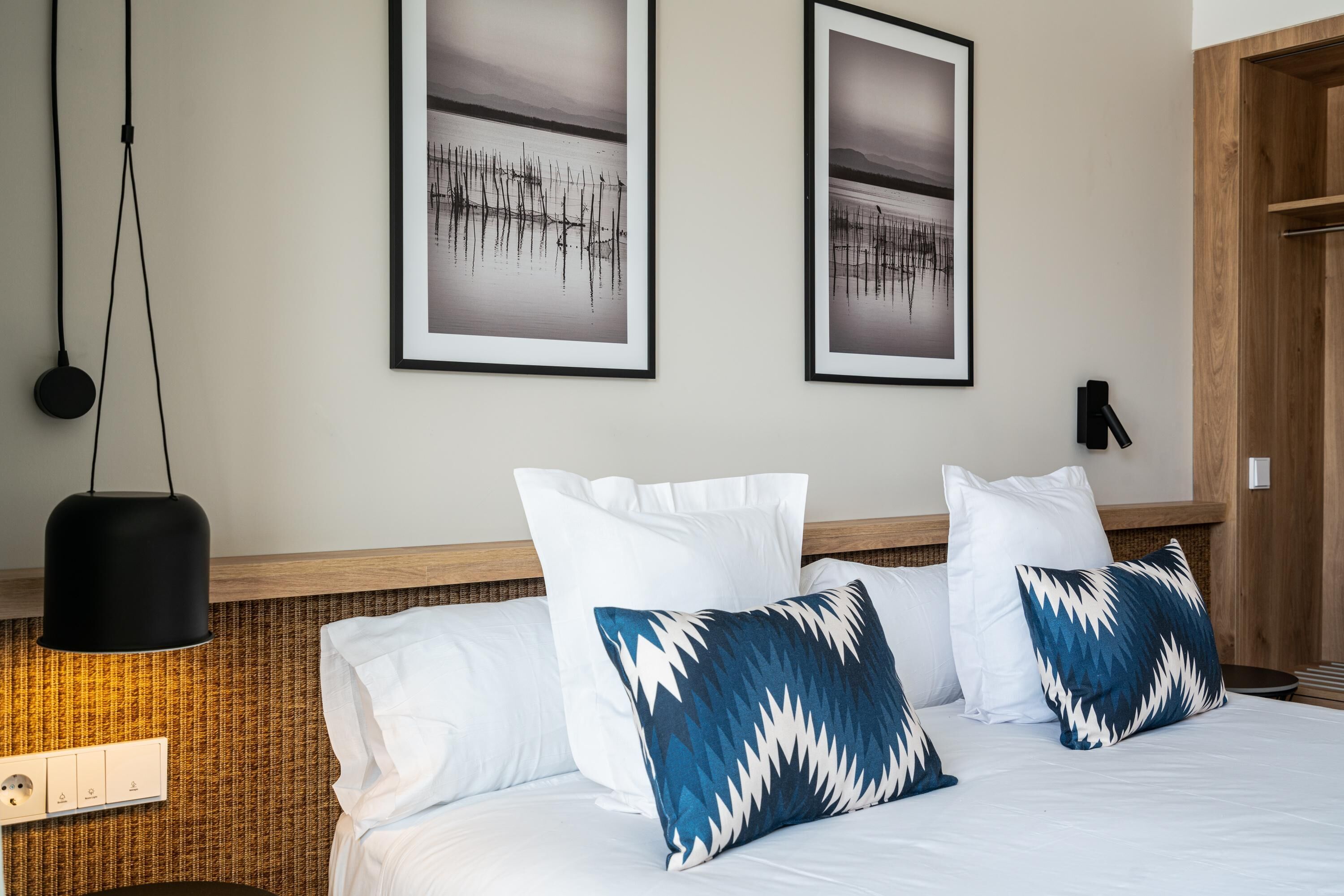 A hotel room with a bed, two framed pictures on the wall, a hanging light, and a wooden headboard.