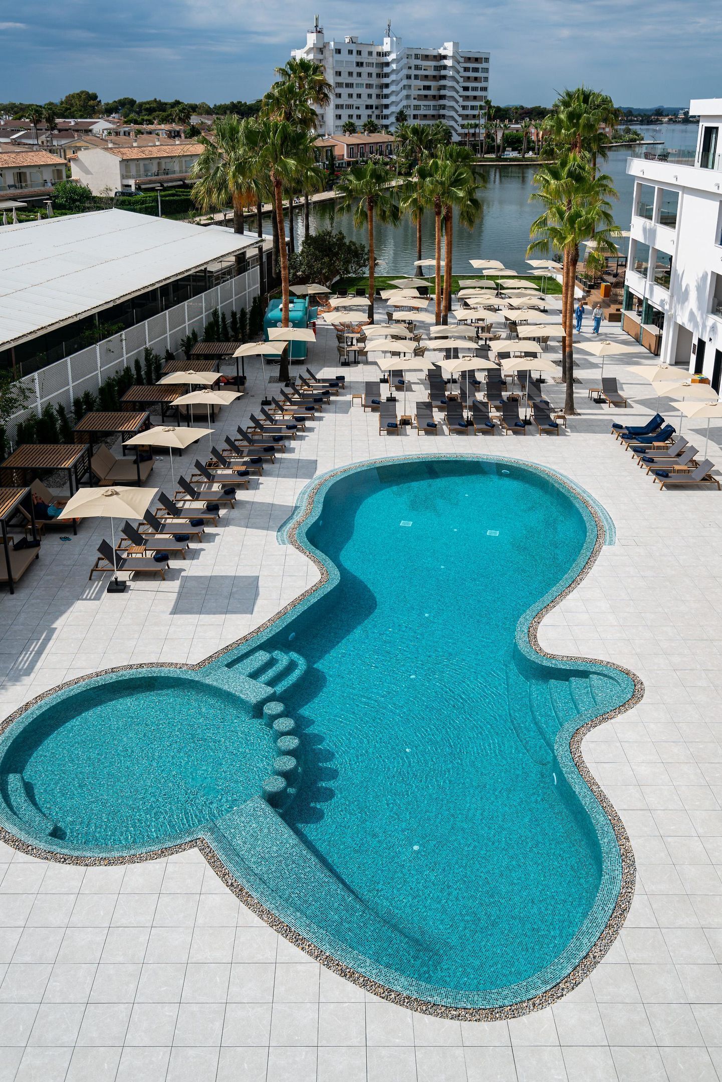 Una piscina con forma singular, rodeada de sillas de descanso y sombrillas, con vista a una masa de agua y edificios al fondo.
