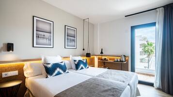 Habitación de hotel con una cama grande, dos cuadros en la pared, una mesita de noche con lámpara y vista al exterior a través de una ventana.