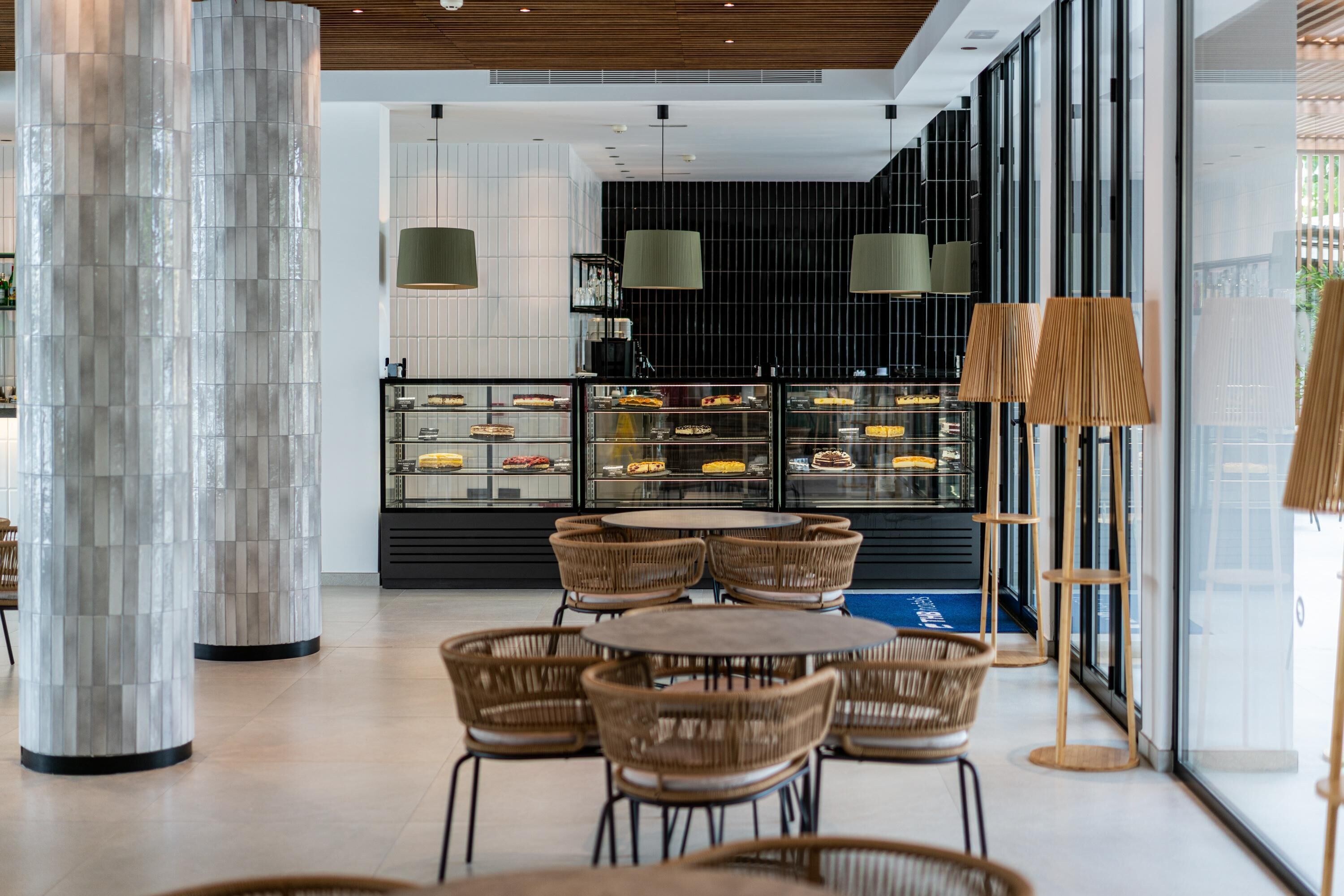 A modern interior with round tables and wicker chairs, a glass display case with pastries, and a large pillar.
