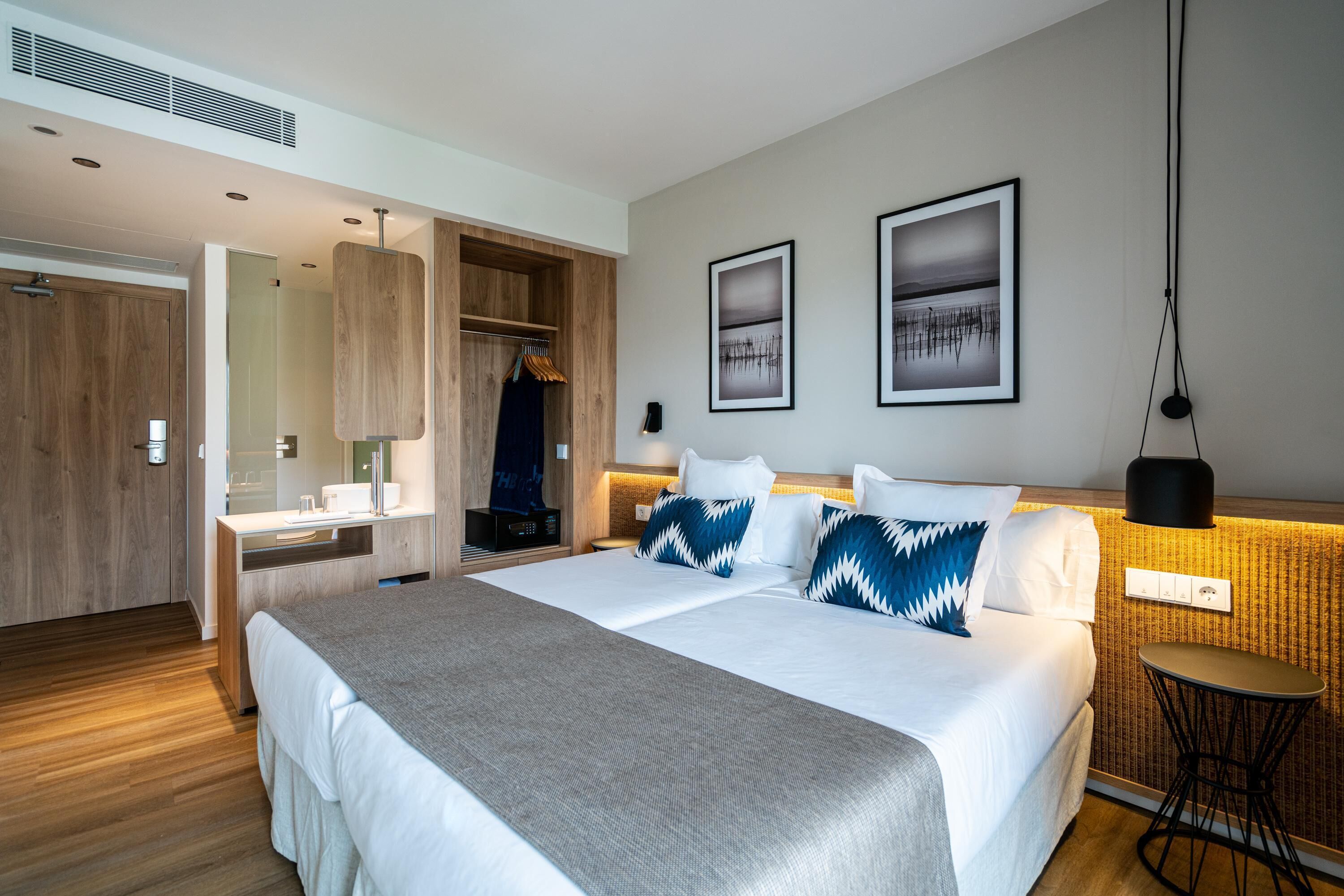 Una habitación de hotel moderna con una cama grande, un armario de madera, una cómoda con espejo y dos cuadros en la pared.