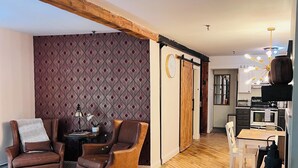 Ein gemütliches Wohnzimmer mit zwei braunen Ledersesseln, Tapete mit Muster, einem Holzbalken an der Decke, einer Schiebetür im Bauernhausstil und einer Küchenzeile mit Herd und Schränken.