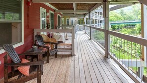 A covered porch with wooden flooring and a railing, featuring outdoor seating and a view of a bridge and greenery.