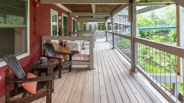 Eine überdachte Veranda mit Holzboden und Geländer, ausgestattet mit Sitzgelegenheiten im Freien und Blick auf eine Brücke und Grünflächen.