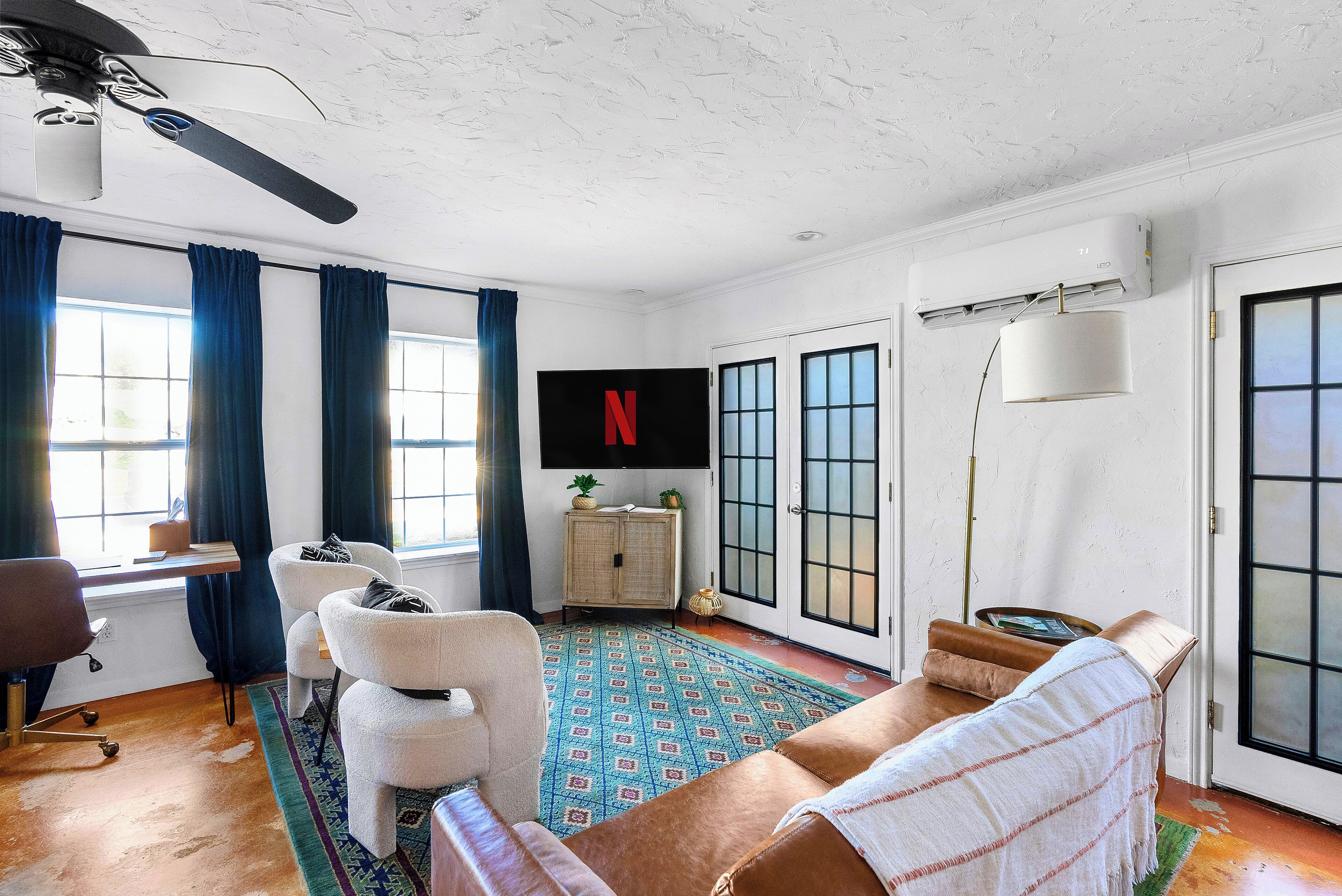 Ein Wohnzimmer mit Deckenventilator, Flachbildfernseher, großem Fenster mit Vorhängen, einer Couch, einem Sessel, einem Schrank und einem gemusterten Teppich.