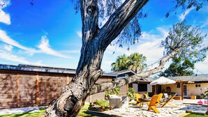 Un patio trasero con un árbol grande, asientos exteriores y un porche cubierto.