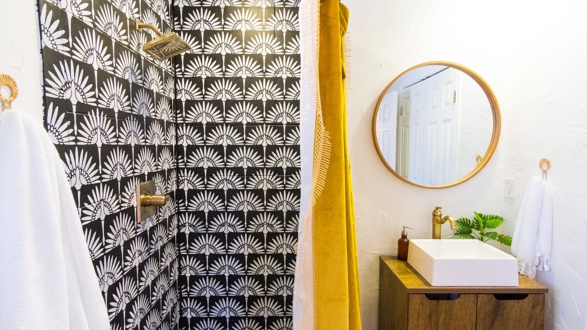 Un bagno con tenda da doccia floreale in bianco e nero, uno specchio rotondo, un mobile bagno in legno con lavandino bianco e un asciugamano giallo.