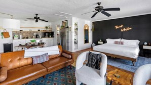 Una habitación moderna y bien iluminada con una cama, un sofá de cuero marrón, un sillón blanco y un comedor con una cocina al fondo.