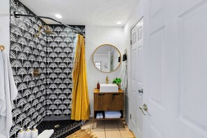 Een badkamer met een douchewand met zwart-wit patroon, een geel douchegordijn, een houten wastafelmeubel en een ronde spiegel.