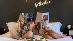 Dos personas sentadas en una cama con una bandeja de tazas, leyendo libros.