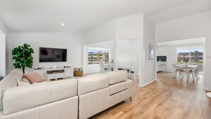 Uma sala de estar ampla com um sofá seccional grande, uma TV de tela plana e um espaço de jantar com mobiliário branco.