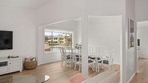 Uma sala de estar espaçosa com área de jantar, janelas amplas com vista para um bairro residencial e uma TV de tela plana na parede.