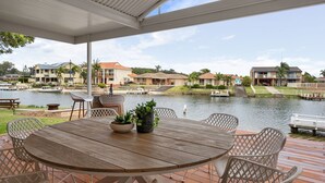 Um alpendre coberto com uma mesa e cadeiras de madeira, com vista para um lago tranquilo com barcos e casas ao fundo.