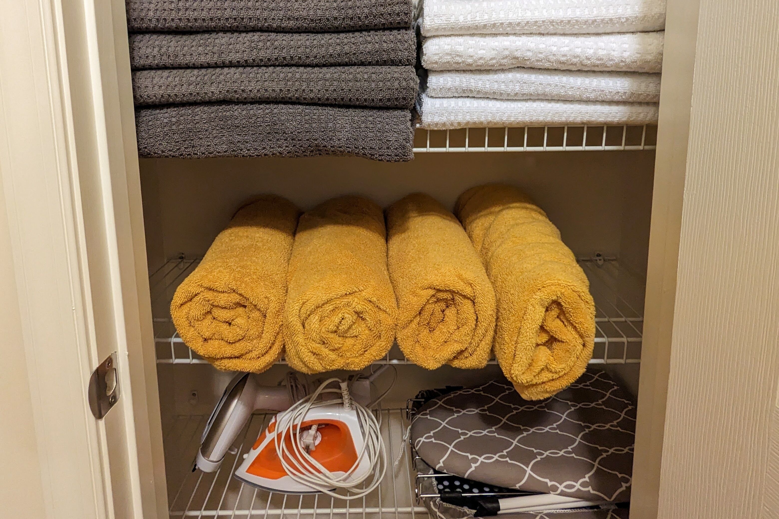 A storage area with neatly folded towels in various colors, a portable iron, and a patterned bag.